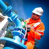 Worker in safety gear adjusting blue industrial piping under clear blue sky, showcasing professional industrial photography by Duncan MacBrayne.