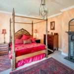 Elegant bedroom featuring a four-poster bed with red bedding, decorative pillows, and warm-toned walls, complemented by a vintage dresser and ornate rug, showcasing the inviting atmosphere captured by Duncan MacBrayne Photography.