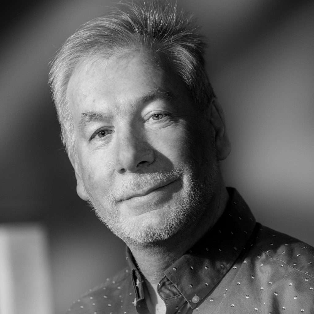 Headshot of Duncan MacBrayne, experienced commercial photographer in Berkshire, with a warm smile and grey hair, wearing a dotted shirt, captured in black and white.