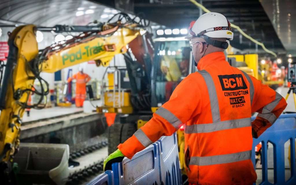 Construction worker in high-visibility orange jacket observing excavation equipment at a railway site, highlighting industrial photography by Duncan MacBrayne in Reading, Berkshire.