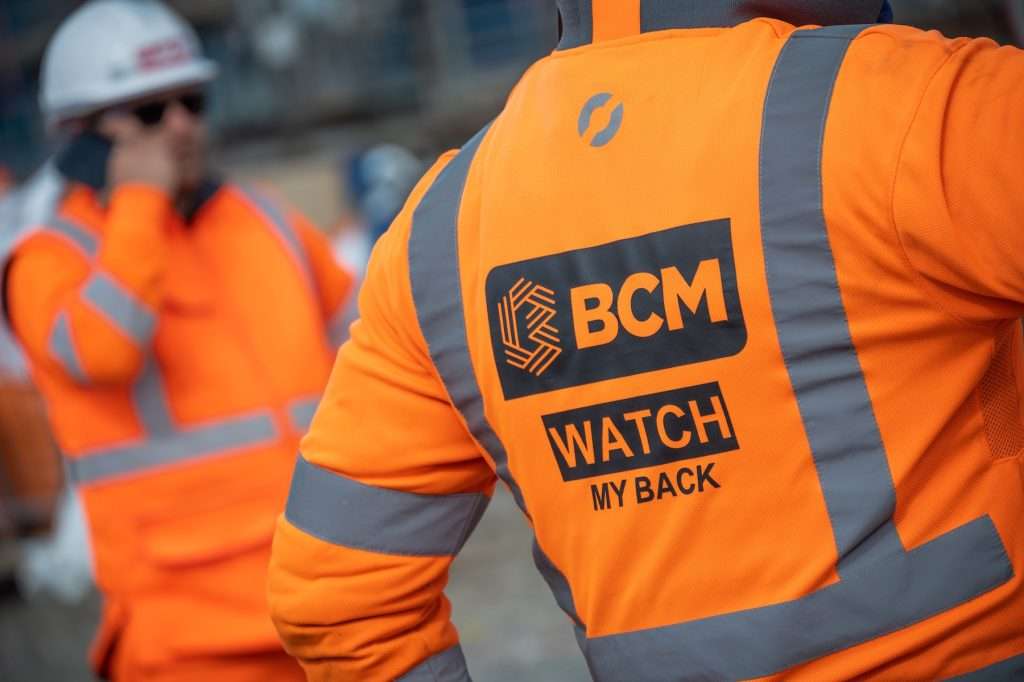 Construction worker in high-visibility orange vest with "BCM WATCH MY BACK" text, focused on safety in an industrial setting.
