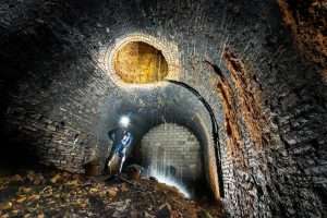 Man standing in a dark, industrial tunnel with brick walls, illuminated by a headlamp, showcasing the interior of a construction site related to the Elizabeth Line project.