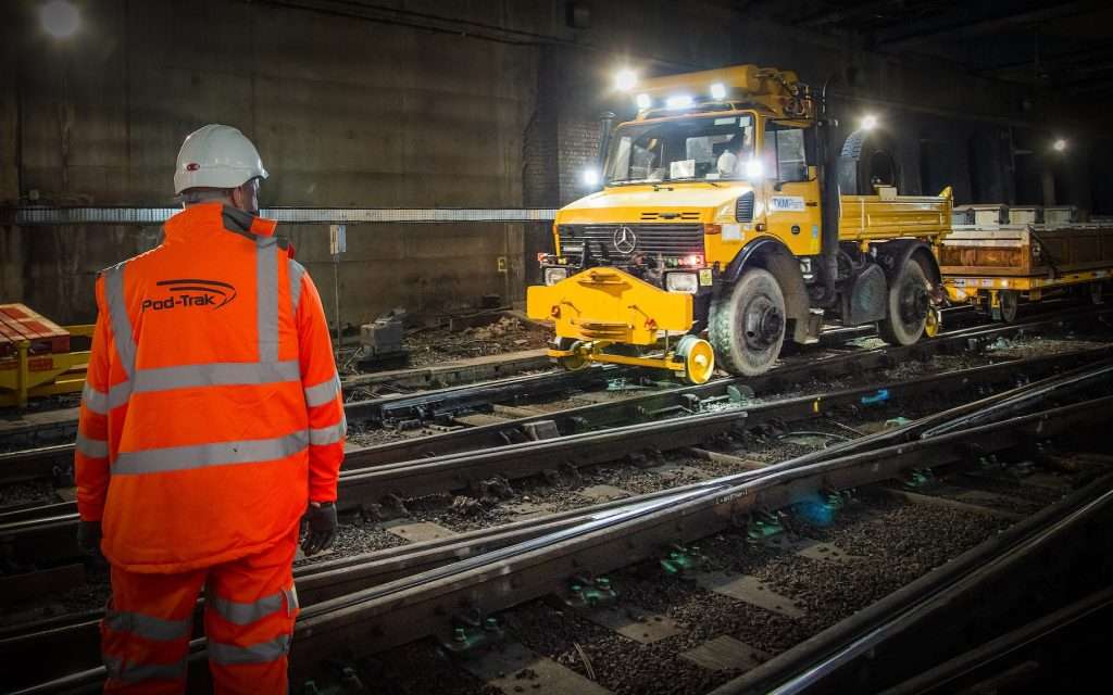 Worker in high-visibility Pod-Trak uniform observing yellow maintenance vehicle on railway tracks in dimly lit industrial setting.