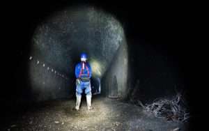Person in safety gear standing in a dark tunnel, illuminated by a flashlight, showcasing industrial exploration and inspection photography by Duncan MacBrayne.