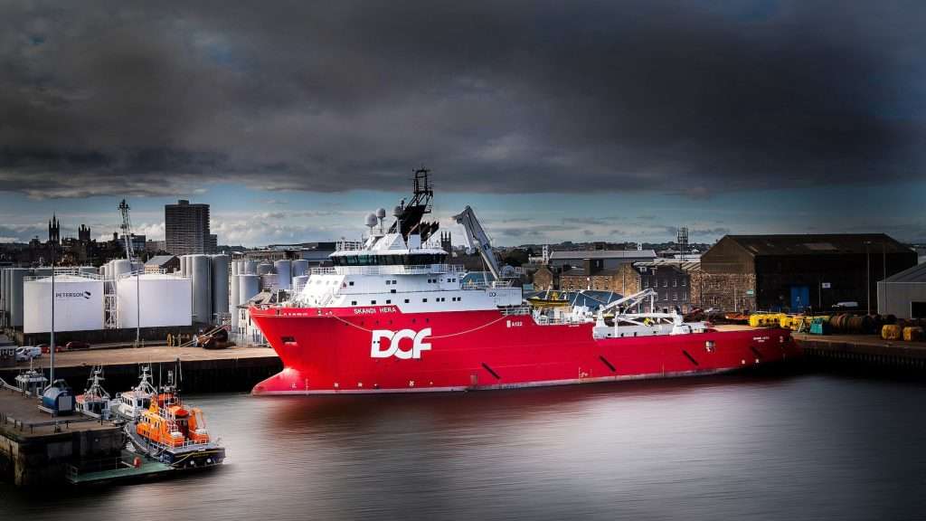 Red industrial support vessel named "Skandi Hera" docked at a port, with city skyline and storage tanks in the background, showcasing marine photography by Duncan MacBrayne.