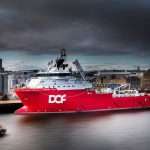 Red industrial support vessel named "DOF" docked in a harbour, with a cloudy sky and cityscape in the background, illustrating marine photography by Duncan MacBrayne.