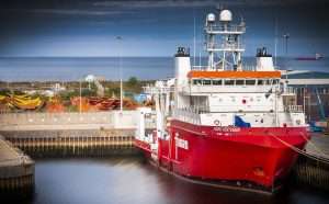 Red industrial research vessel docked in harbour with fishing boats in the background, showcasing Duncan MacBrayne's commercial photography expertise in marine settings.