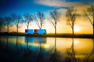 Truck parked beside a tranquil river at sunset, silhouetted by trees, reflecting vibrant colours in the water, showcasing Duncan MacBrayne's commercial photography.