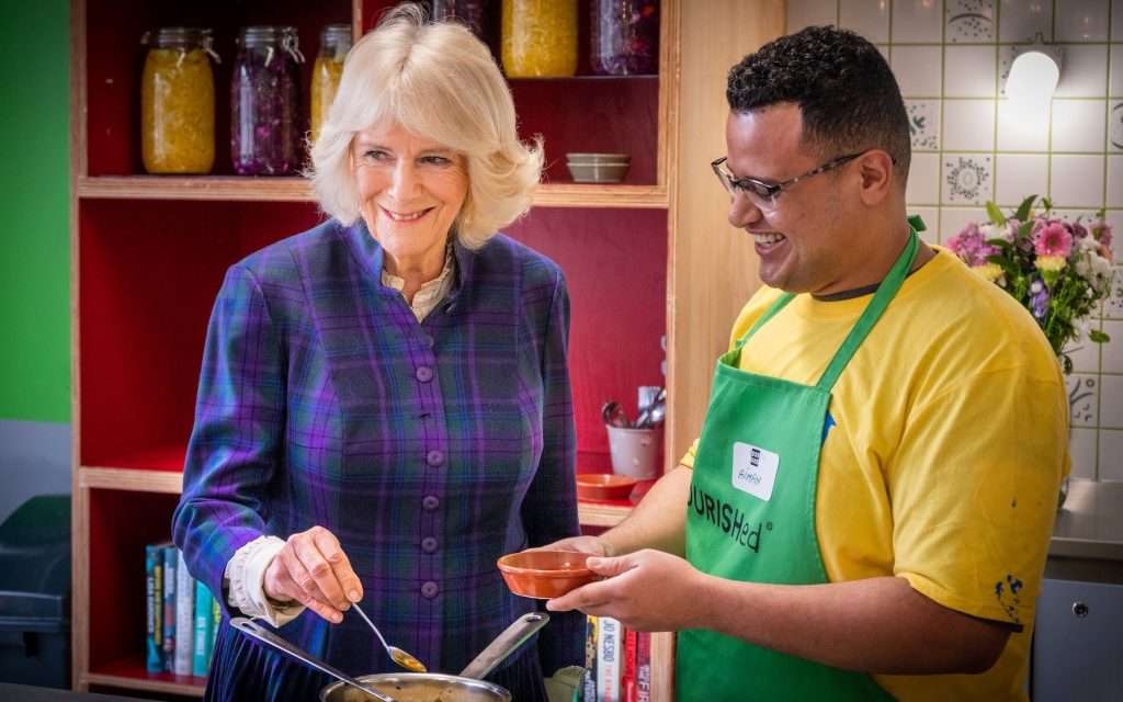 Queen Camilla in a tartan dress smiling while stirring a pot, interacting with a man in a green apron holding a bowl, in a vibrant kitchen setting with shelves of jars and books.