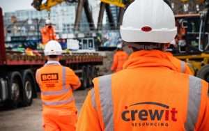 Construction workers in high-visibility orange jackets and hard hats, featuring 'crewit resourcing' logo, overseeing machinery at a site, representing industrial photography services by Duncan MacBrayne.