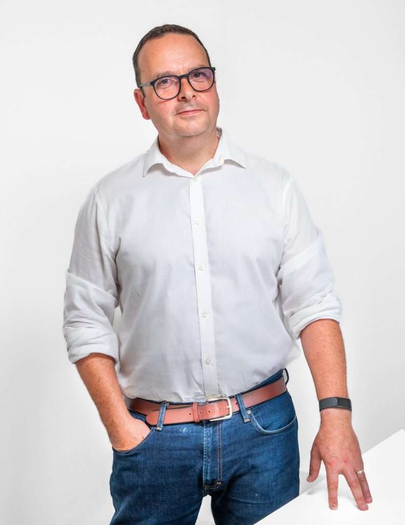 Professional portrait of a man in a white shirt and blue jeans, posing confidently with one hand on his hip, showcasing Duncan MacBrayne's commercial photography style.