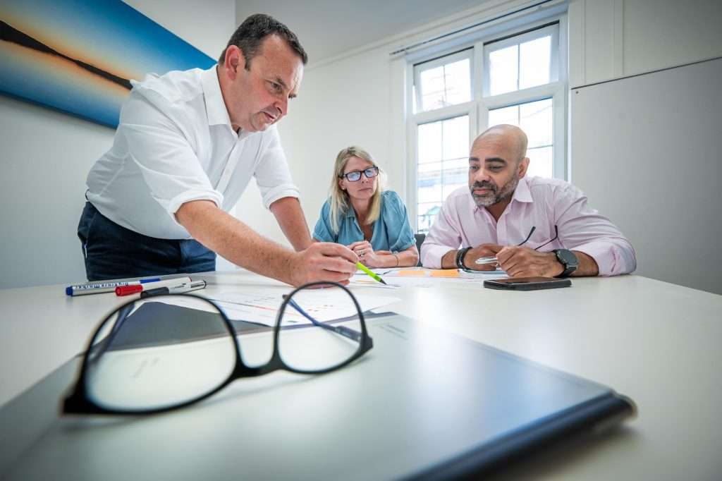 Business meeting with three professionals discussing plans at a table, featuring glasses and stationery, in a bright office environment.