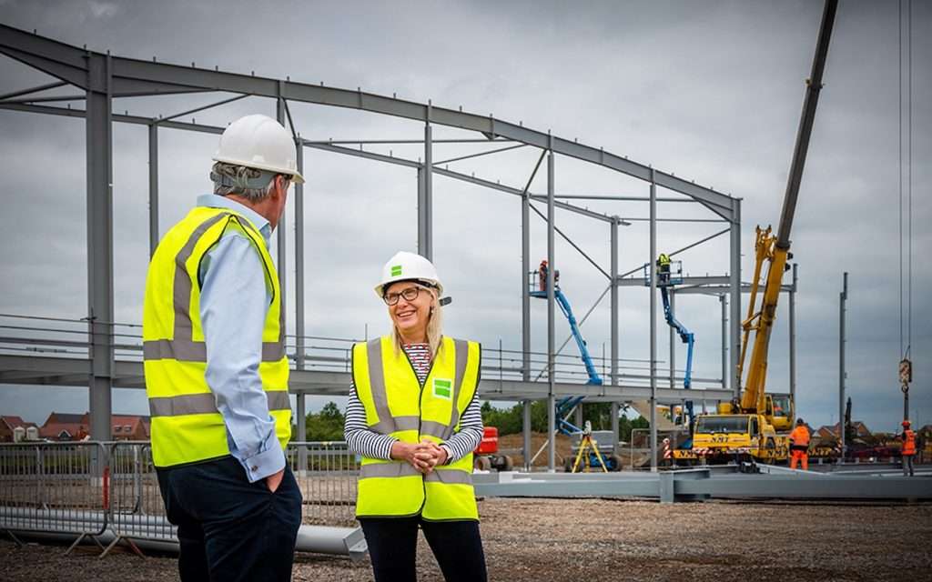 Construction site with two professionals in hard hats and high-visibility vests discussing project details, steel framework in background, showcasing industrial photography by Duncan MacBrayne.