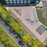 Aerial view of a commercial property parking lot with solar panels, surrounded by greenery and adjacent to a road, showcasing industrial and commercial photography by Duncan MacBrayne.