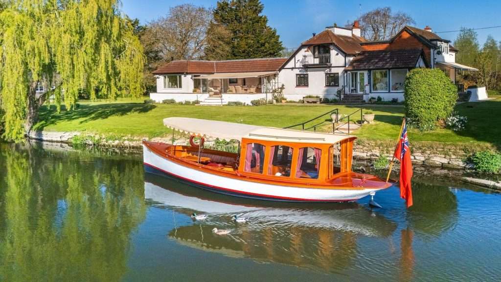 Boathouse on a riverbank with a wooden boat in the foreground, surrounded by lush greenery and a residential property in Reading, Berkshire, showcasing luxury property photography.