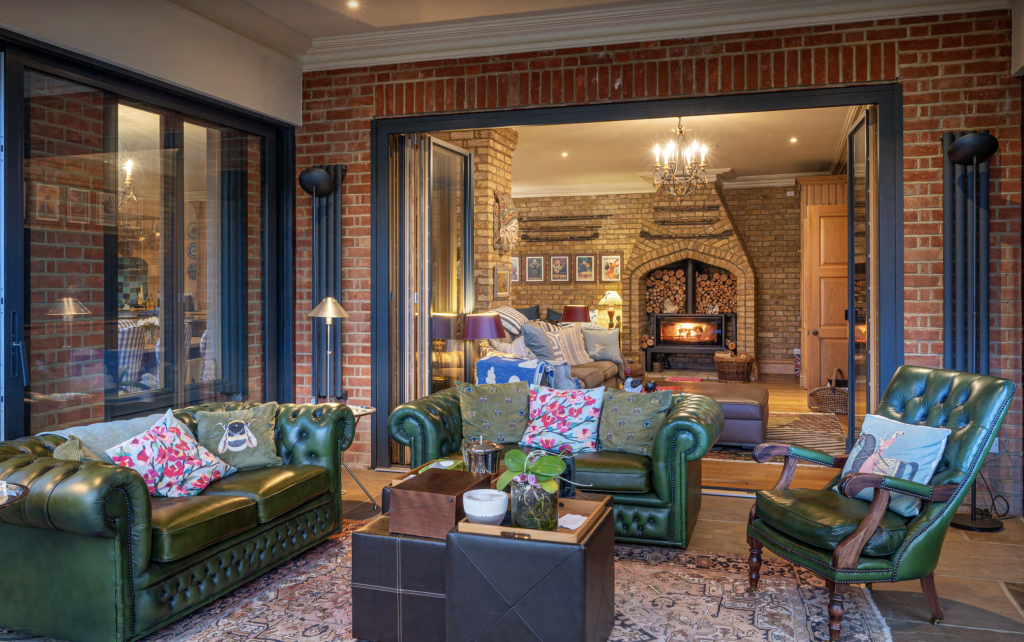 Duncan MacBrayne Luxurey Residential Property Luxurious green leather sofas with floral cushions in a stylish living room featuring a warm fireplace, brick walls, and elegant decor, showcasing high-quality interior photography for luxury property marketing.